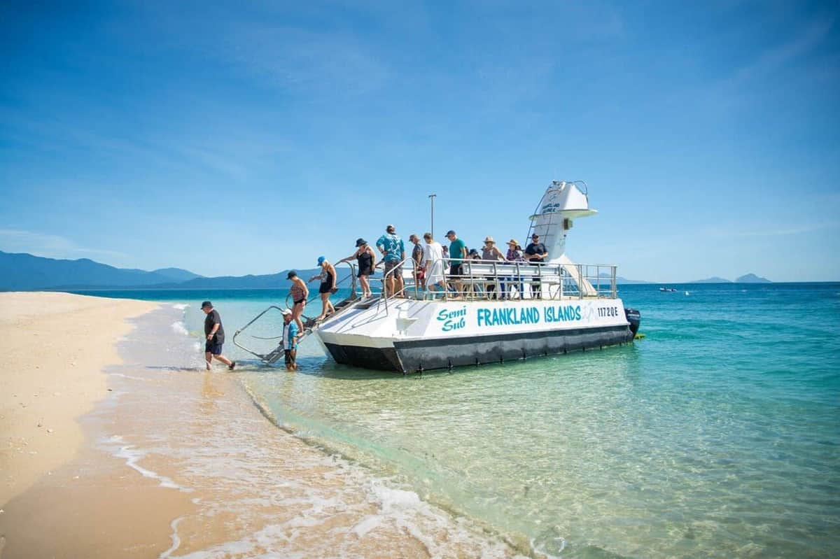 Frankland Islands Reef: Boat Tour from Cairns A group of people disembark from a boat labeled "FRANKLAND ISLANDS" onto a sandy beach with clear, blue water under a sunny sky.