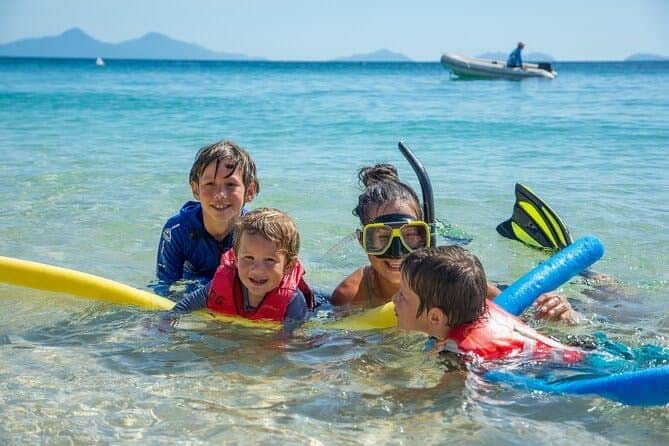 Frankland Islands Reef: Guided Day Cruise from Cairns + Snorkeling + Lunch Four children in life vests playing in shallow ocean water, one wearing a snorkel mask. Mountains and a boat are in the background.
