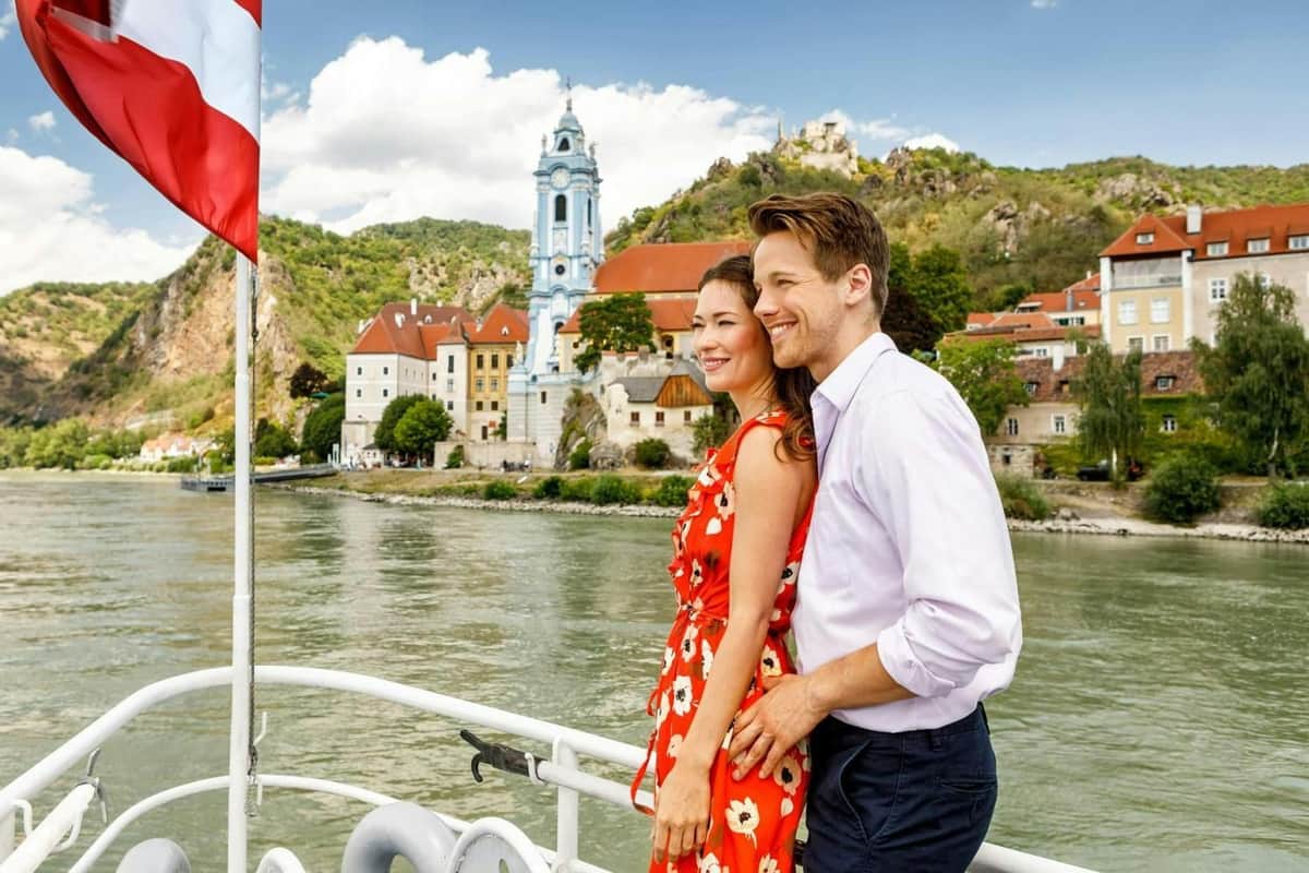Danube Valley: Day Trip from Vienna A smiling couple stands on a boat with a scenic riverside town with colorful buildings and a blue church tower in the background.