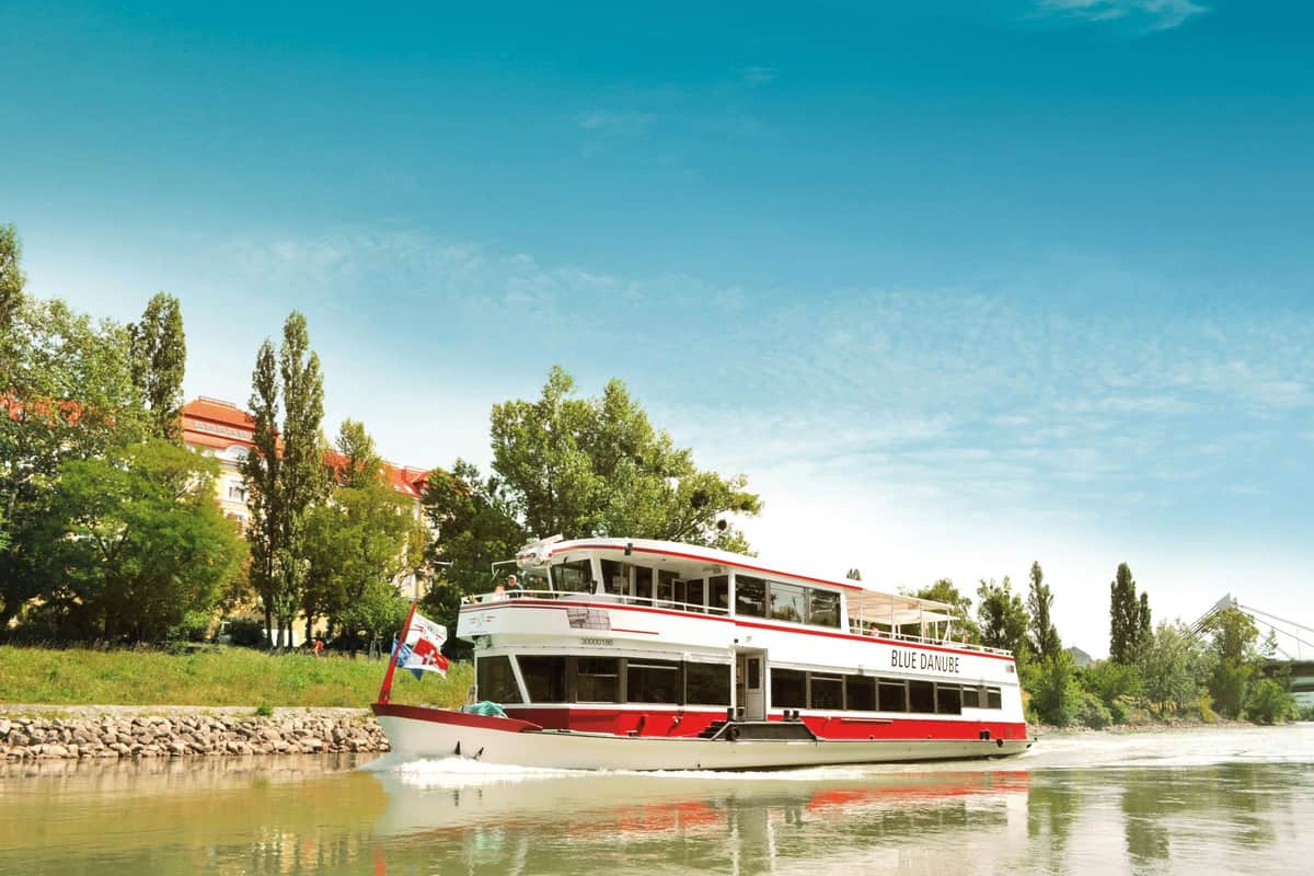 Vienne : Croisière touristique Un bateau fluvial nommé "Danube bleu" navigue sur un fleuve près d'un rivage avec des arbres et des bâtiments, sous un ciel bleu clair.