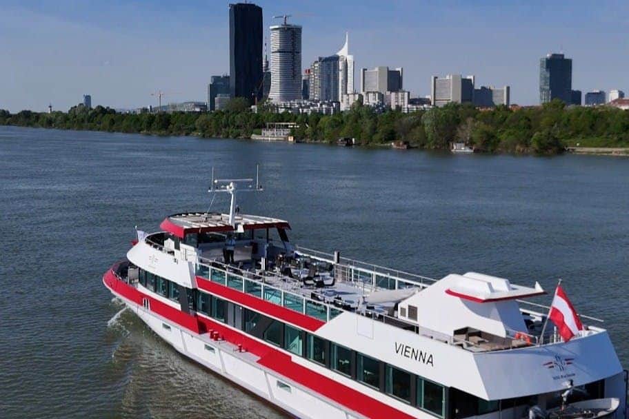 Vienna: Danube Cruise A boat named "Vienna" travels on a river with a cityscape featuring tall buildings in the background and trees lining the riverbank.