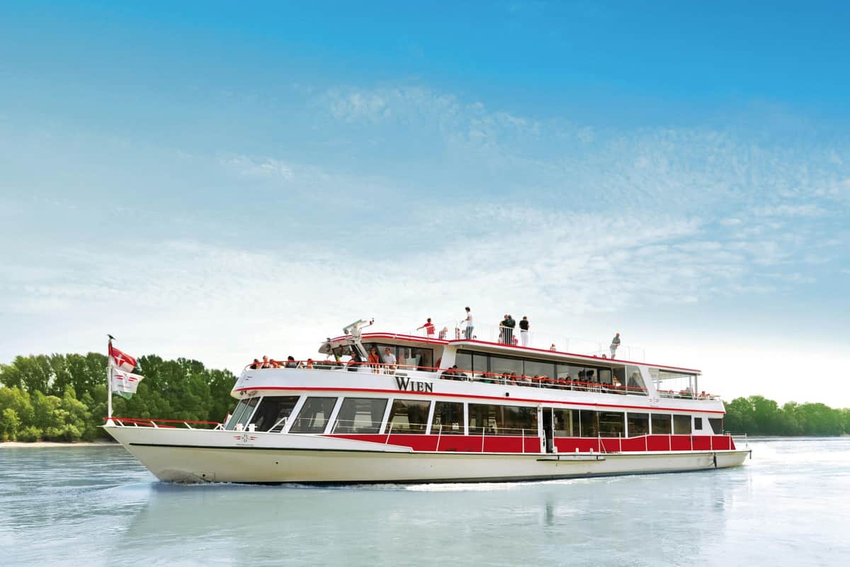 Vienna: Grand Danube River Cruise A white and red passenger boat named "Wien" with people on the upper deck sails on a body of water under a clear blue sky.