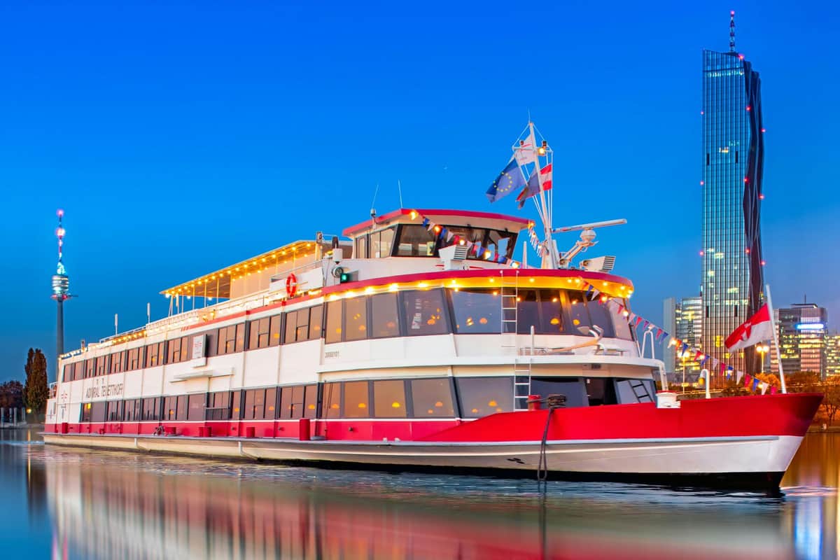 Vienne : Croisière Heurigen avec chansons viennoises + buffet Un bateau blanc et rouge décoré de guirlandes lumineuses et de drapeaux, amarré dans un port sur fond de ciel bleu clair.