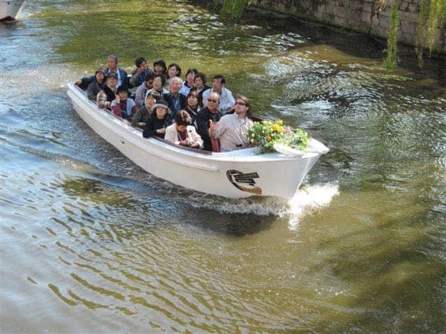 Historical Boat Tour Including 1 Ghent Beer A group of people seated in a white boat cruising along a canal. A floral arrangement is at the front of the boat.