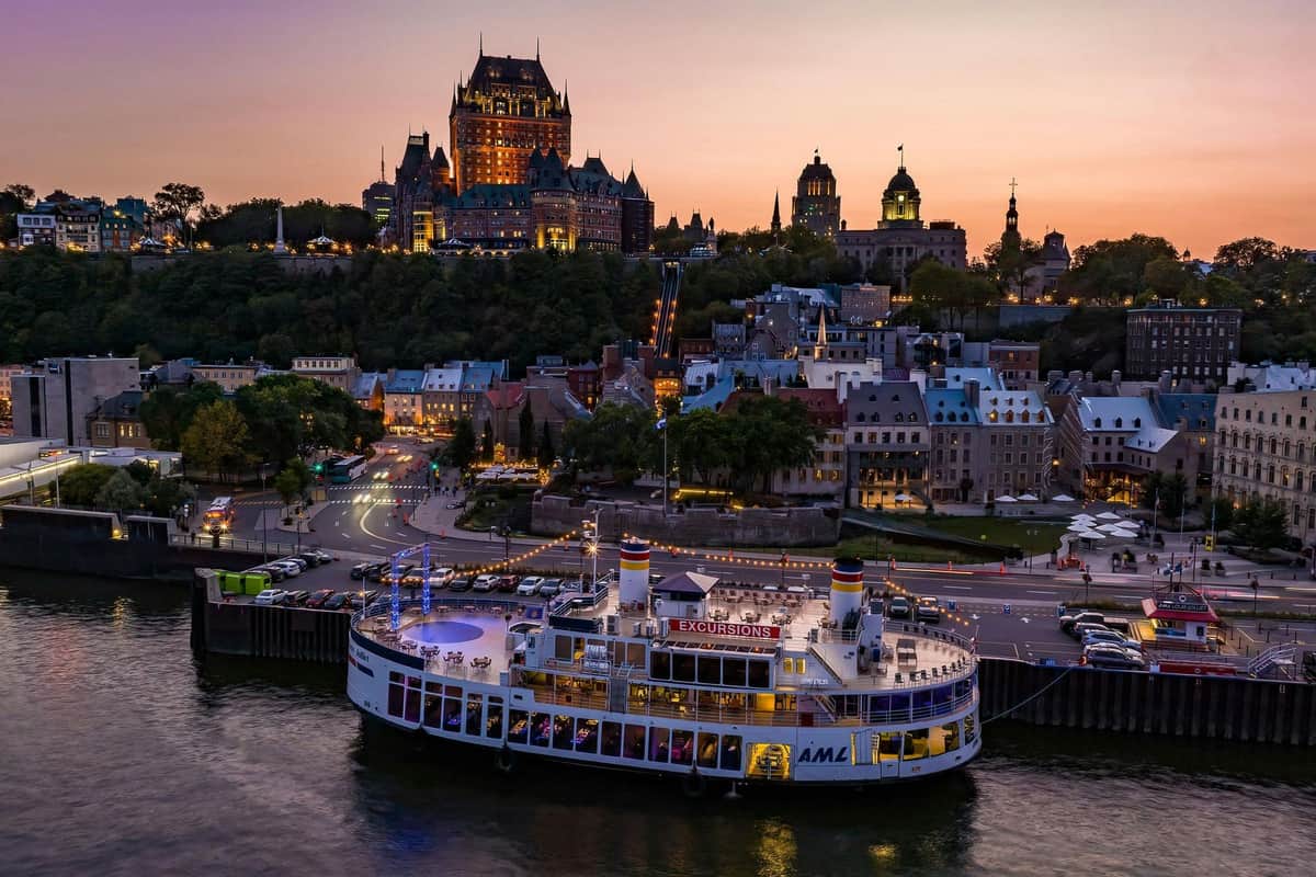 Québec : Croisière en soirée avec DJ Un ferry illuminé à quai sur une rivière avec en arrière-plan un paysage urbain historique et des bâtiments illuminés sur une colline au crépuscule.