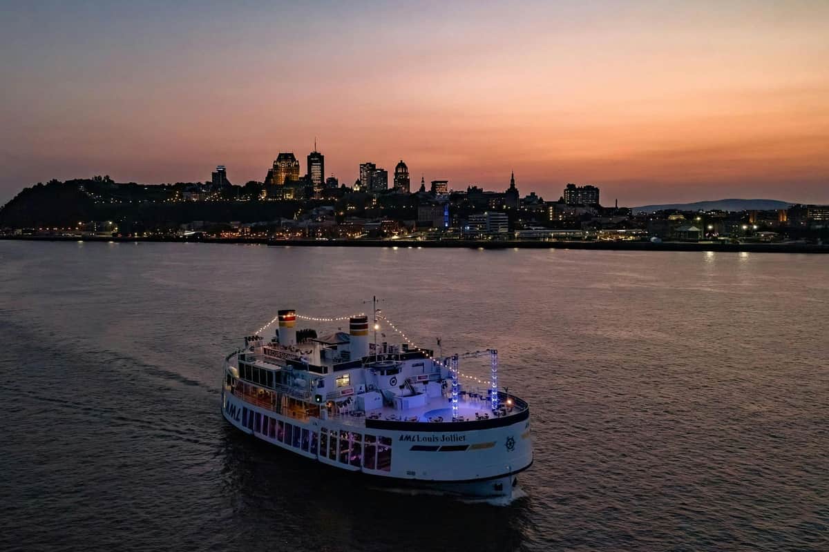 Québec : Croisière fluviale guidée en soirée Un ferry illuminé naviguant sur une rivière au crépuscule, avec en arrière-plan une ligne d'horizon urbaine composée de bâtiments illuminés.