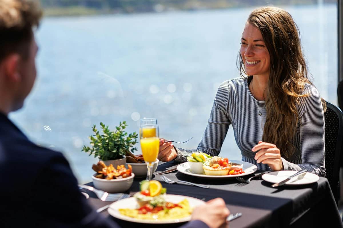 Quebec City: Weekend Sightseeing Brunch Cruise A woman smiling while having a meal at a table with plates of food and a glass of orange juice, overlooking water.
