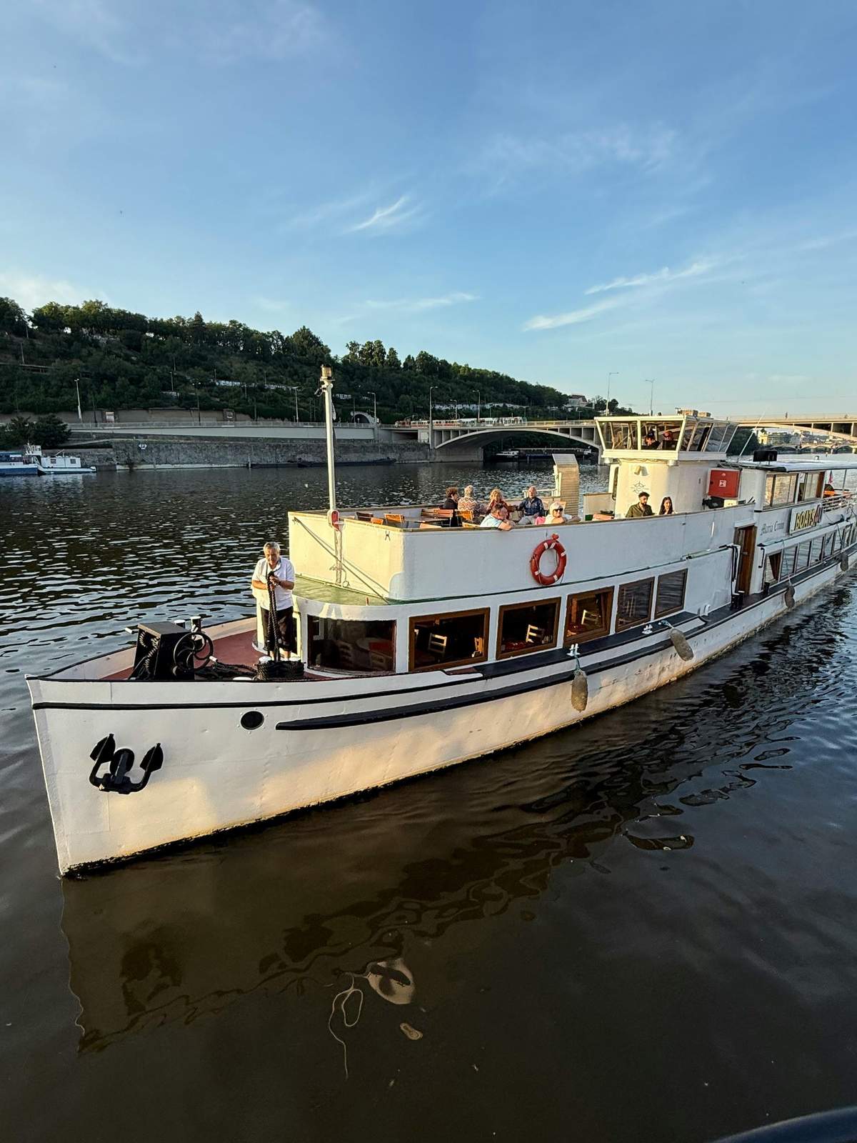 Prague : Visite en bateau + audioguide + collation et boisson Un bateau blanc avec des personnes sur son pont supérieur navigue sur une rivière calme, avec un pont et de la verdure en arrière-plan.