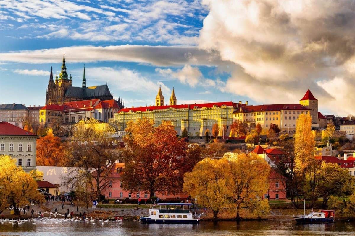 Prague: City Tour with Lunch & River Cruise View of colorful autumn trees, historic buildings, and a church with spires under a partly cloudy sky. A boat is on a river.