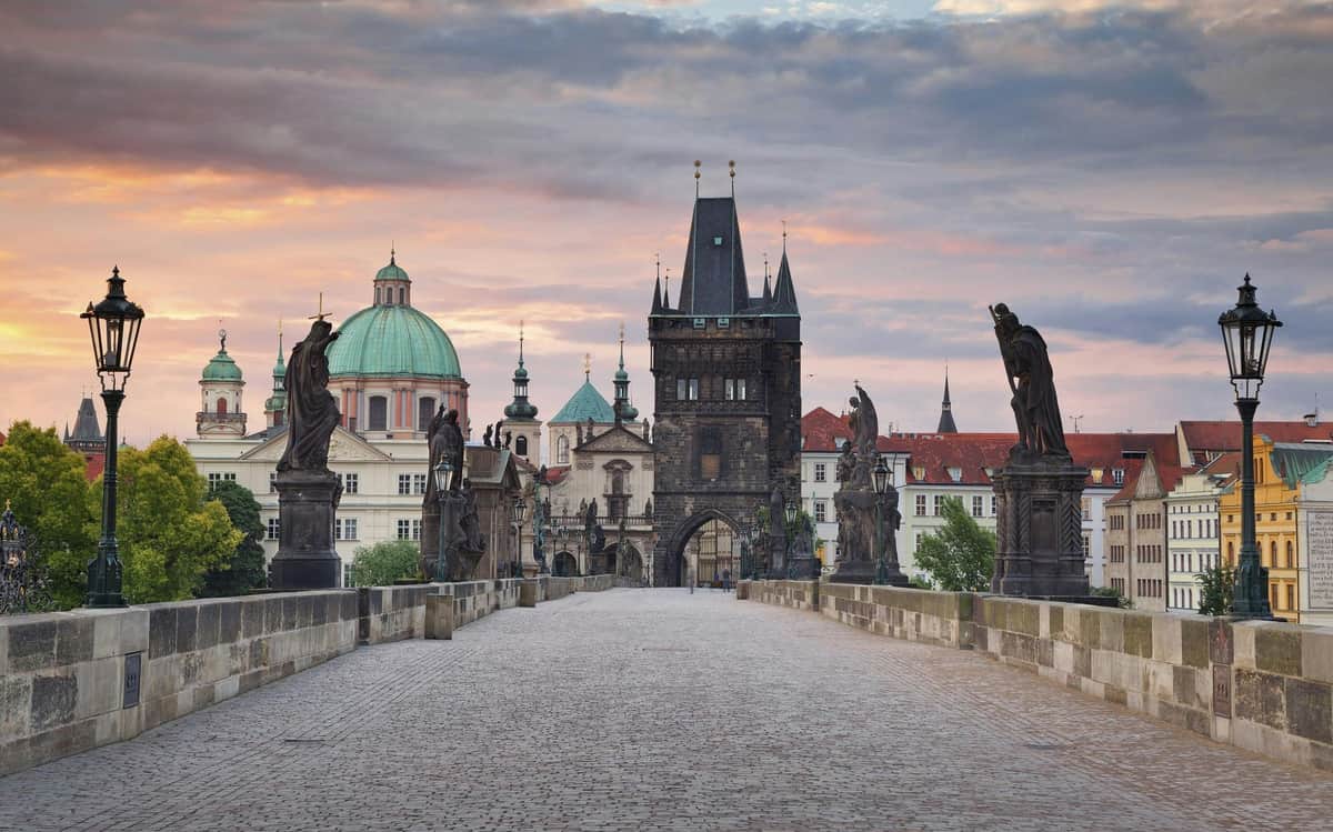 Prague CoolPass : Accès à plus de 80 attractions de premier plan + réductions Pont historique en pierre avec statues, menant à une tour gothique et à des bâtiments en forme de dôme sous un ciel coloré au coucher du soleil.