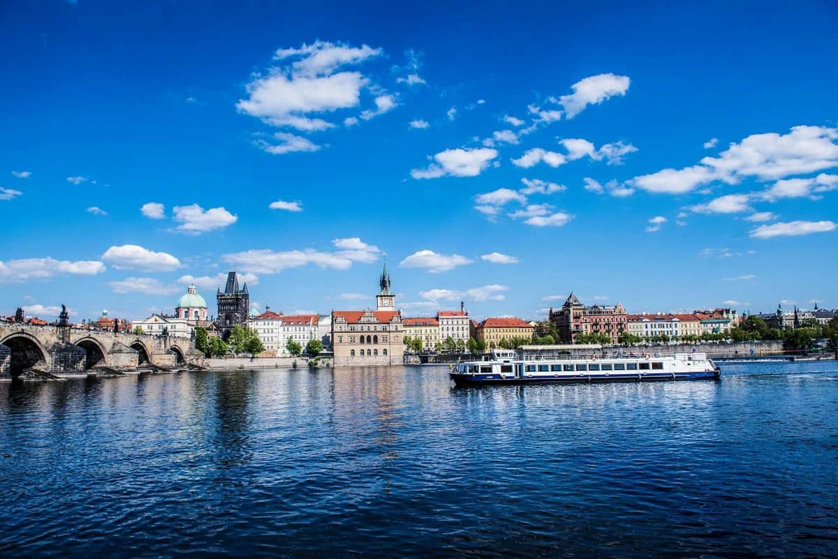 Prague: Sightseeing Cruise + Dinner and Live Music A boat cruises on a river with historic buildings and a clear blue sky with clouds in the background.
