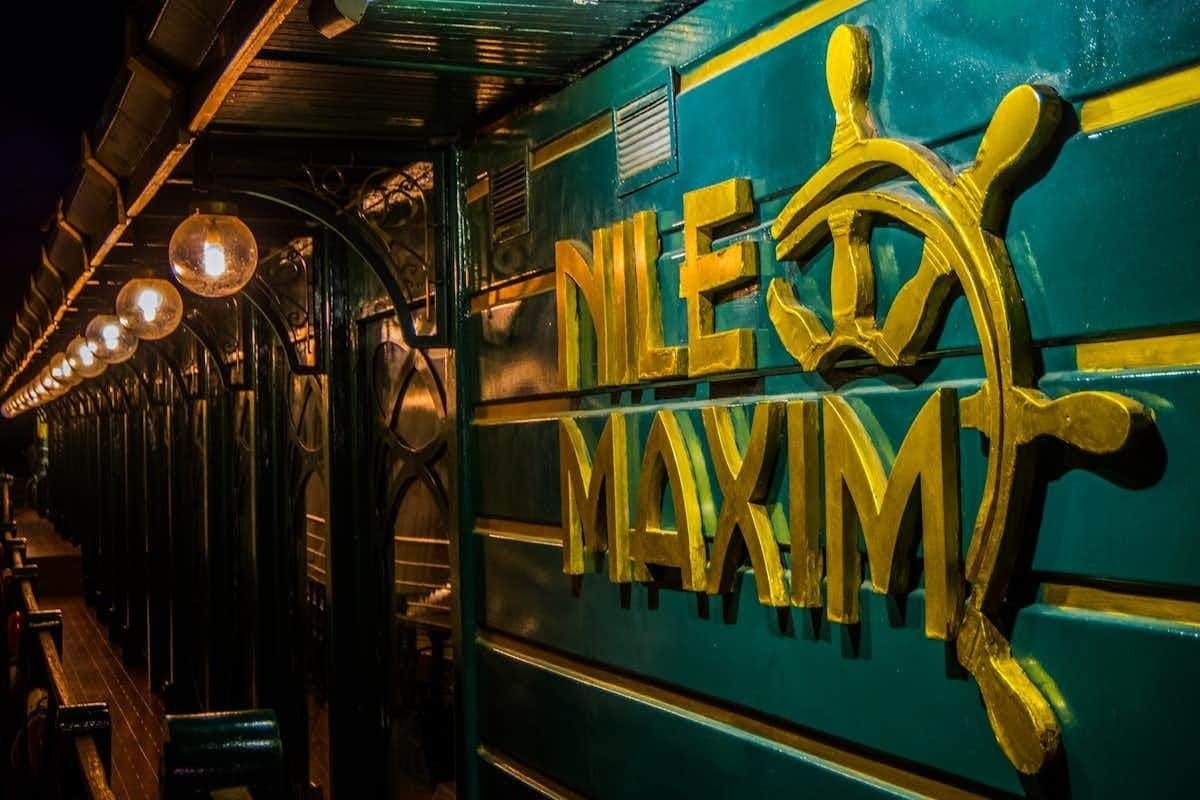 Cairo: Nile Maxim Dinner Cruise + Belly Dance & Tanura + Optional Transfers A teal wall with yellow decorative text reading "Nile Maxim," with ornate black ironwork and warm lighting nearby.