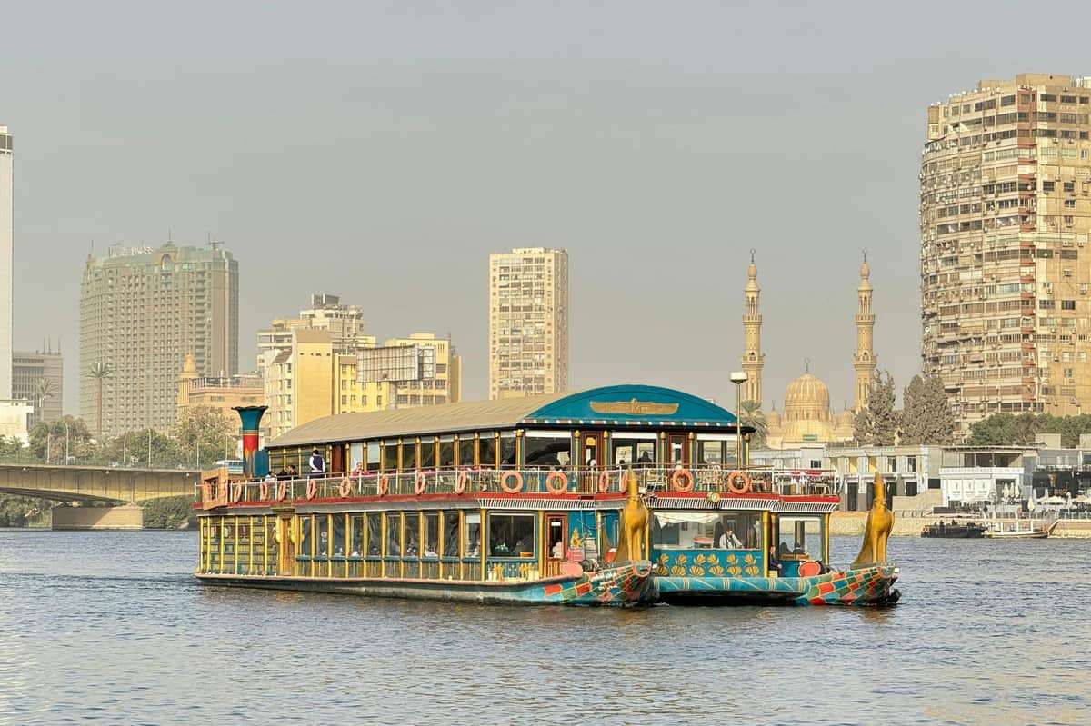 Cairo: Nile Pharaohs Dinner Cruise + Optional Transfer & Drinks A riverboat styled with Egyptian motifs on a river, with a city skyline and two minarets in the background.
