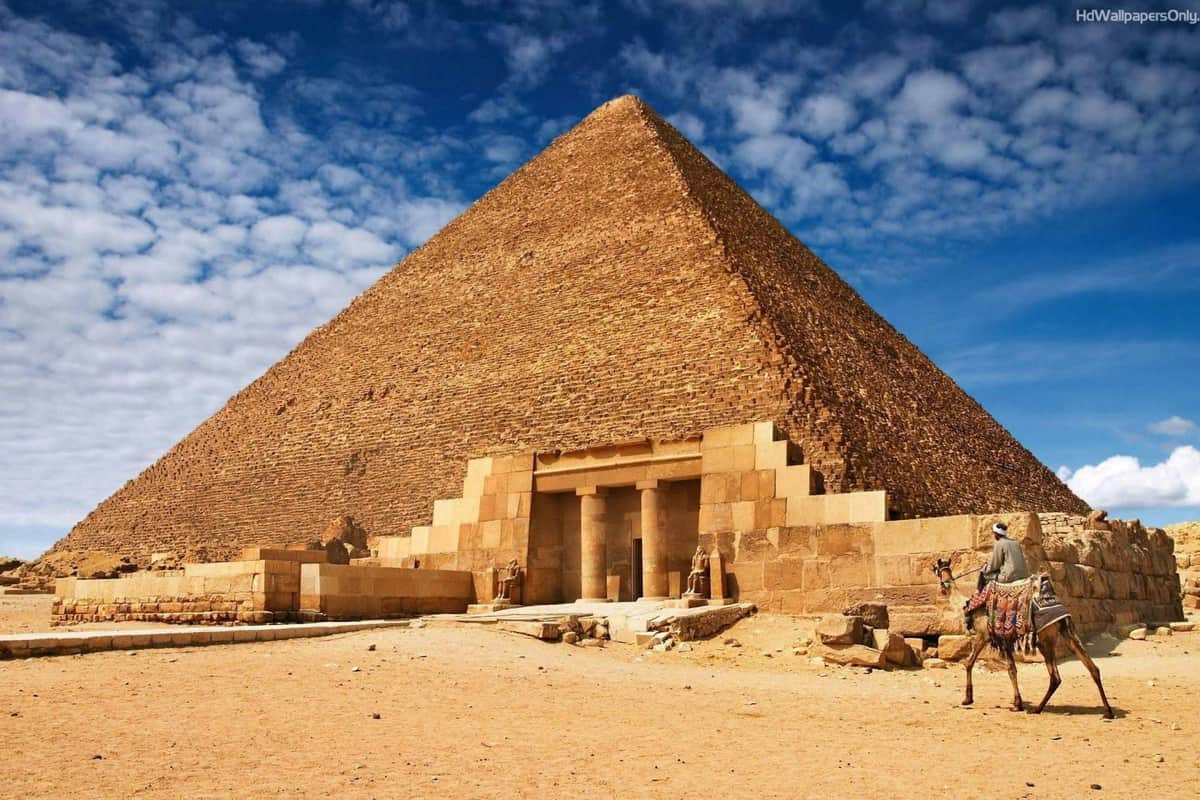 Pyramides de Gizeh : Entrée coupe-file + Dîner-croisière sur le Nil + Transferts privés La Grande Pyramide de Gizeh sous un ciel bleu avec des nuages épars, montrant son entrée et le sol sablonneux qui l'entoure.