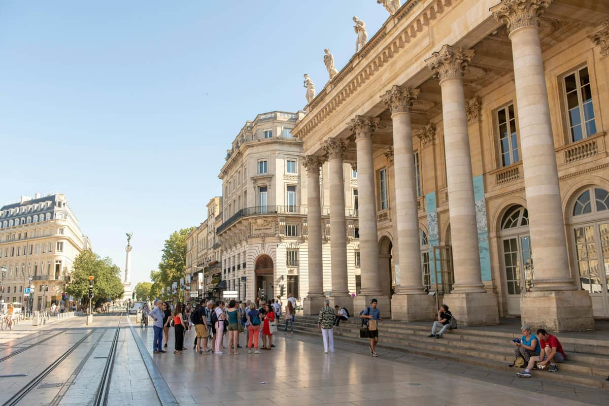 Bordeaux CityPass Un groupe de personnes se tient à l'extérieur d'un grand bâtiment avec des colonnes et des statues sur le toit, sous un ciel bleu clair.