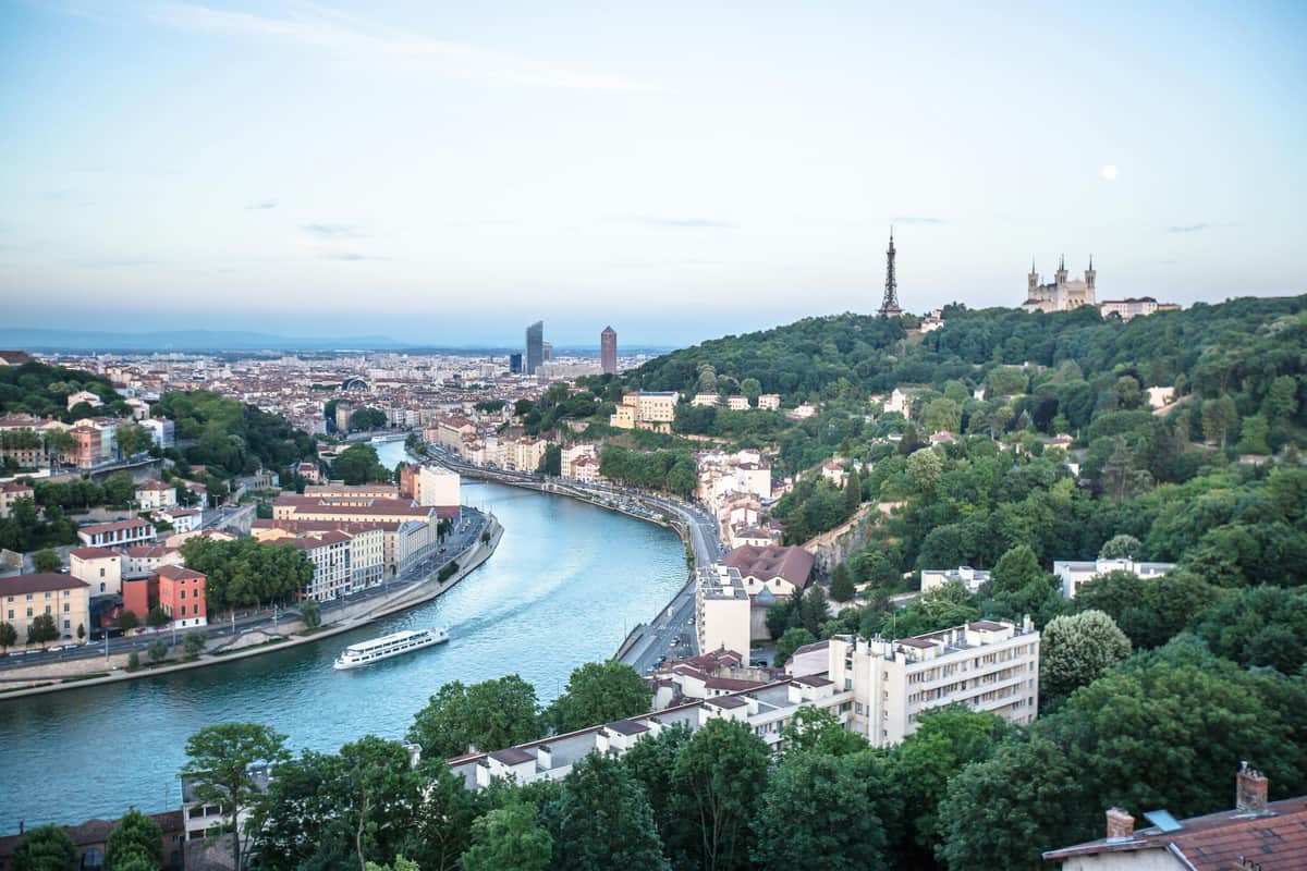 Dinner Cruise on the Saône by Les Bateaux Lyonnais Hermès I A cityscape featuring buildings along a river, a boat cruising on the water, and a hill with a tower and basilica in the distance.