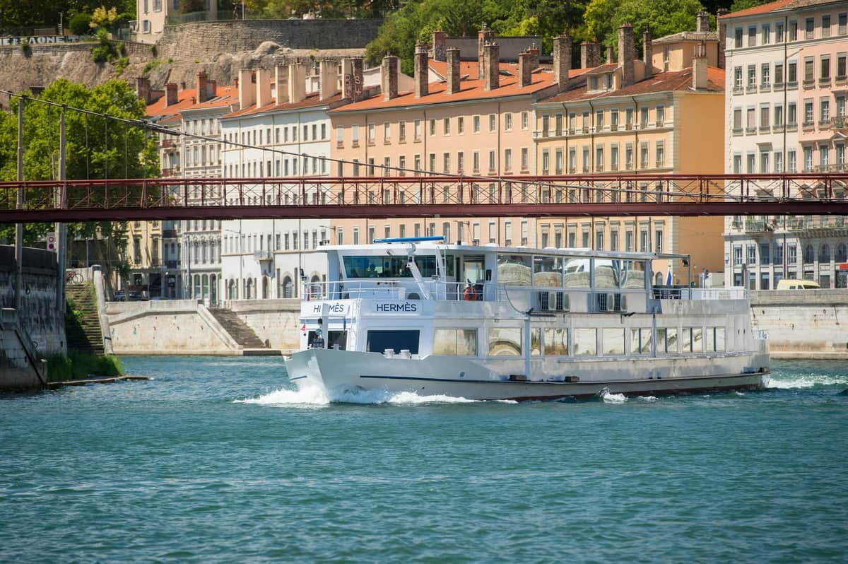 Dinner Cruise on the Saône by Les Bateaux Lyonnais Hermès II A white riverboat named "Hermès" travels on a river, with colorful historic buildings and a red pedestrian bridge in the background.