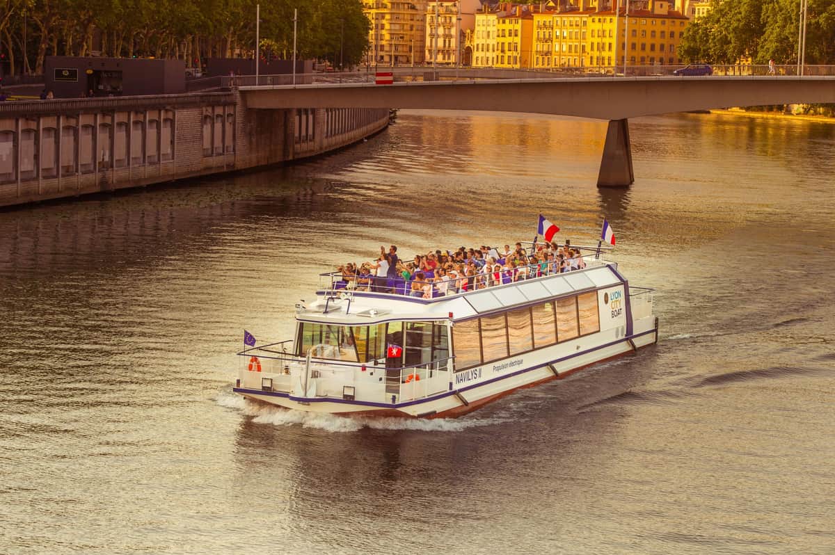 Guided Sightseeing Cruise on the Saône by Les Bateaux Lyonnais A white river cruise boat with passengers, flags, and a bridge in the background on a calm waterway at sunset.