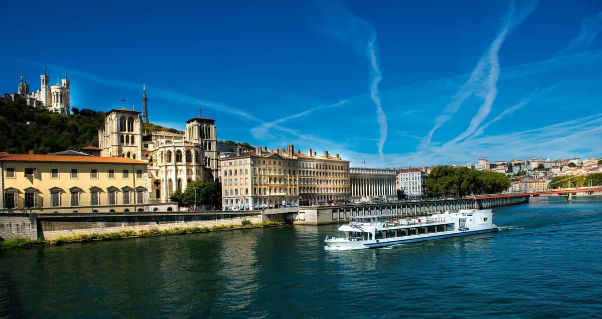 Déjeuner-croisière sur la Saône par Les Bateaux Lyonnais Hermès II Un bateau blanc navigue sur une rivière avec des bâtiments historiques, dont une grande église, le long de la rive opposée, sous un ciel bleu clair.