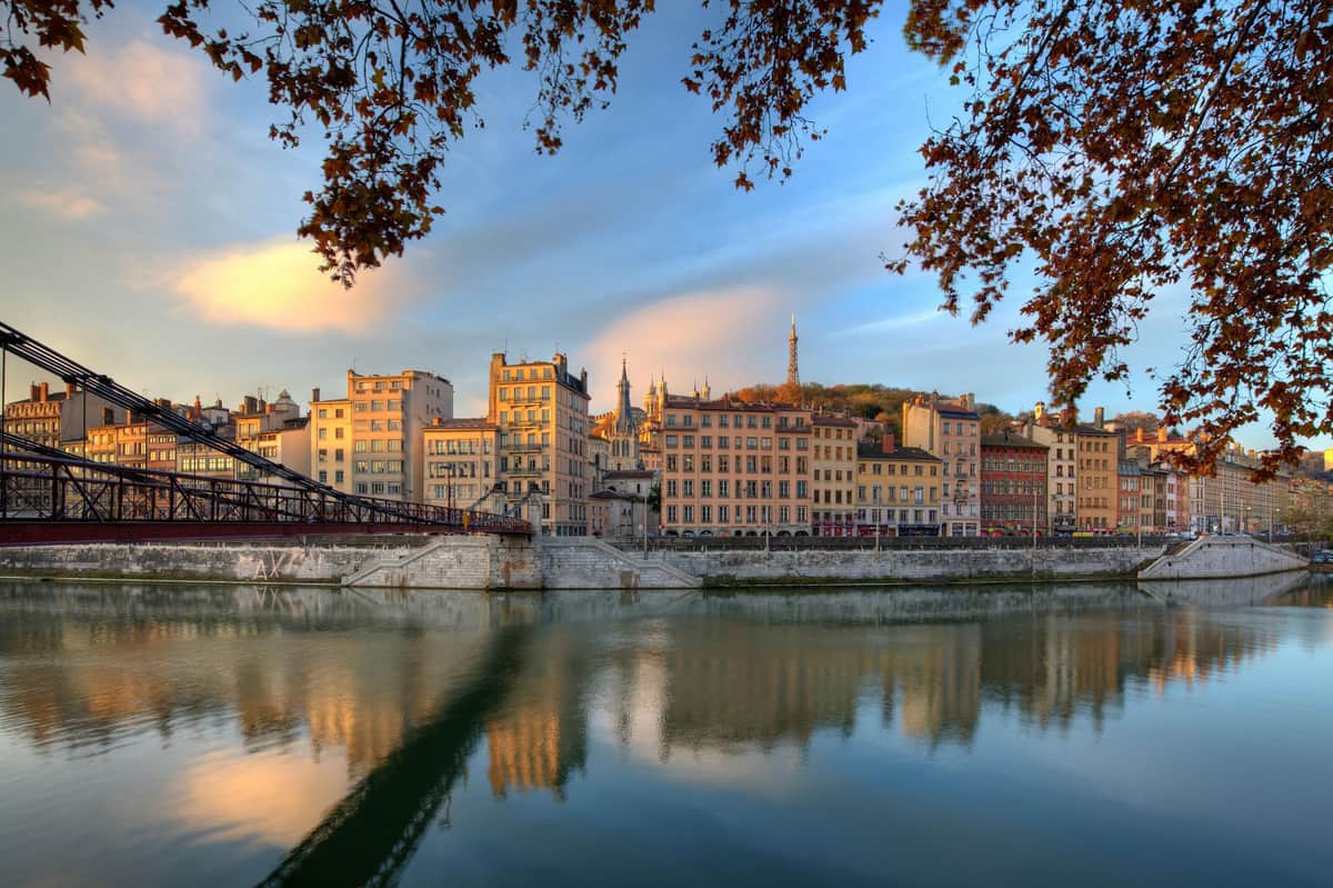 Lyon City Card A riverside view of a city with historic buildings, a pedestrian bridge, and autumn trees under a blue and orange sky.