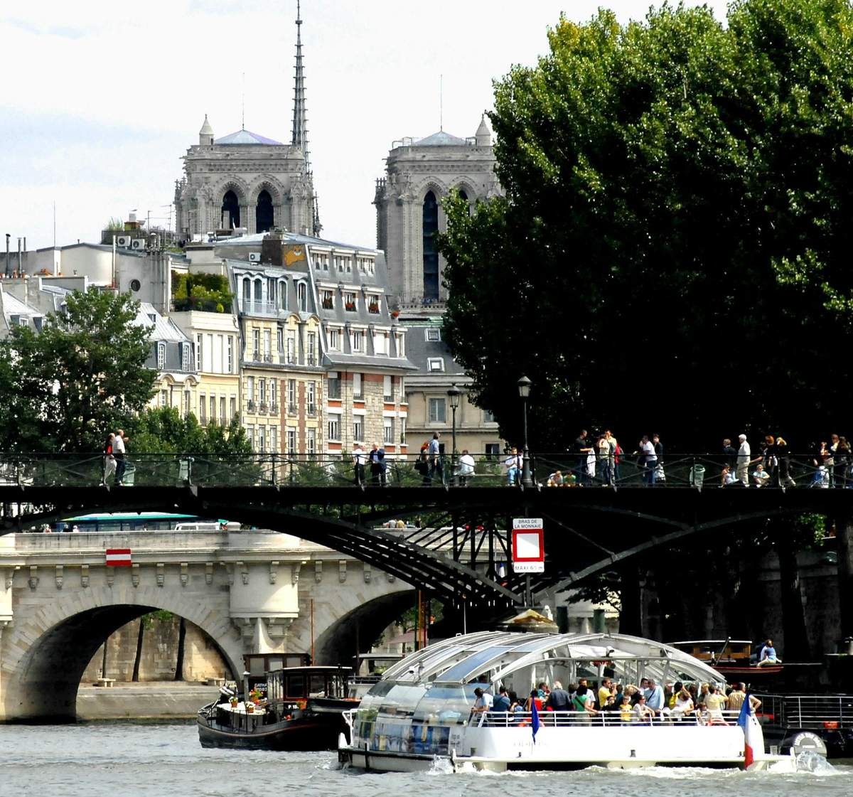 Batobus Paris: Hop-on Hop-off Riverboat People walking on a bridge over a river with boats below, historical buildings, and trees in the background.