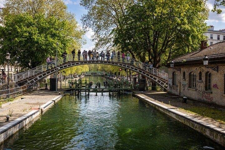 Canal Saint-Martin & Seine River: Guided Sightseeing Cruise A pedestrian bridge crosses a canal surrounded by green trees. People are walking and standing on the bridge.