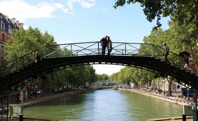 Due persone si trovano su un ponte ad arco su un canale costeggiato da alberi e sentieri in una giornata di sole.