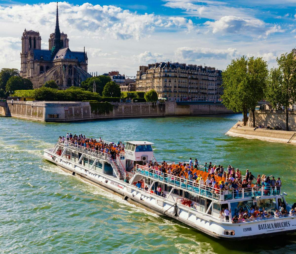 Seine River: Cruise by BATEAUX-MOUCHES® A tourist boat full of people sails on a river with Notre-Dame Cathedral and Parisian buildings in the background.