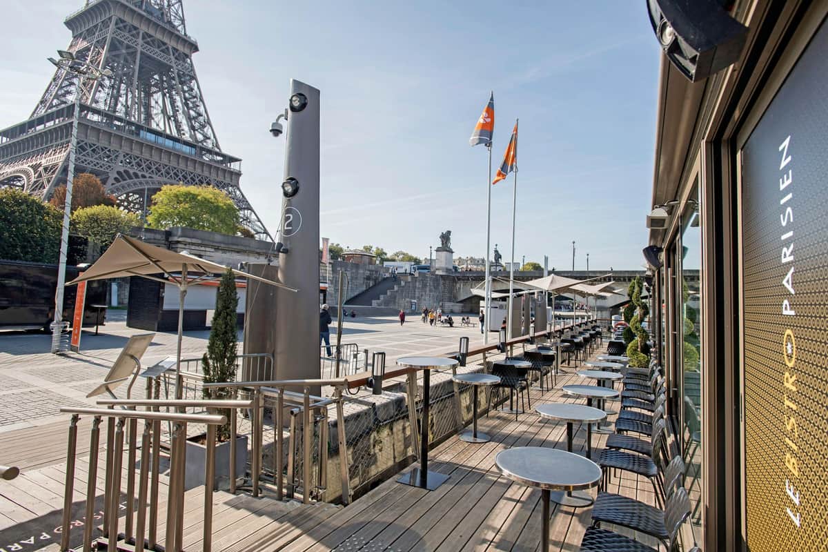 Seine River: Sightseeing Cruise + Lunch at Le Bistro Parisien Outdoor café seating area with empty tables and chairs beside the Eiffel Tower, flags, and few people in the square.