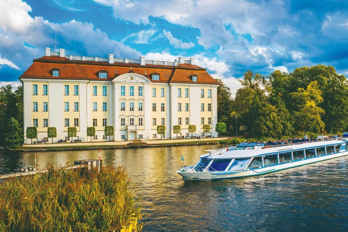 Berlin: 3-Hr Boat Trip to Müggelsee A river with a boat passing in front of a large, yellow, classical-style building under a partly cloudy sky.