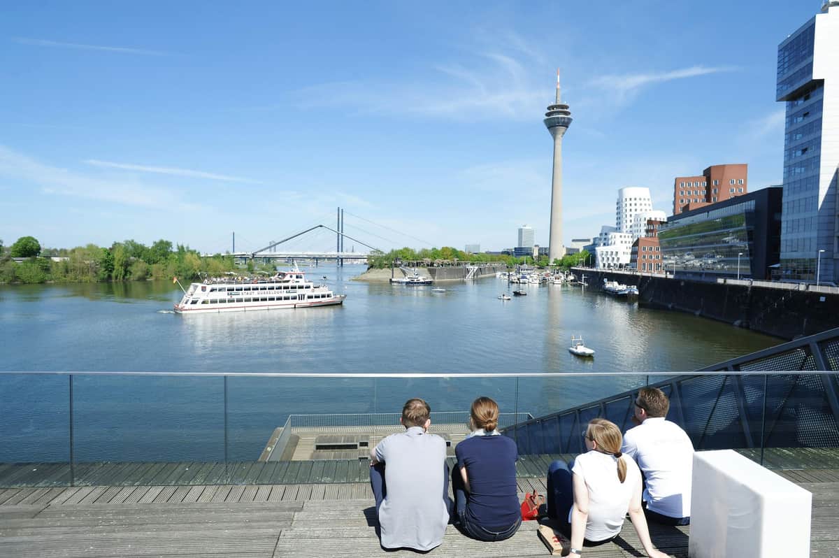 DüsseldorfCard Quatre personnes sont assises sur des marches en bois près d'un port, avec des bateaux, un pont et une haute tour d'observation en arrière-plan.
