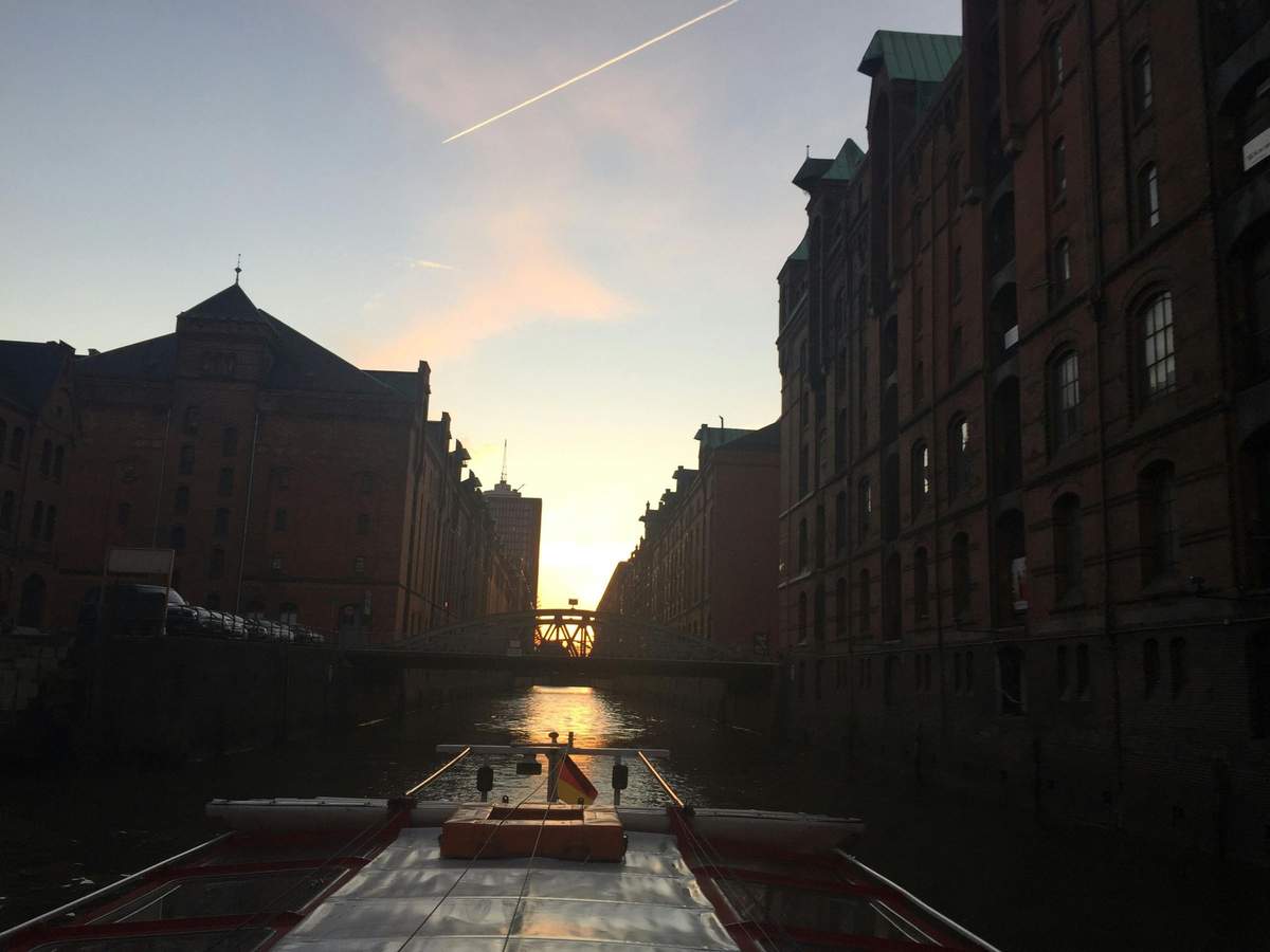 Hamburg: 1.5-Hour Night Cruise Boat sailing between tall brick buildings in a canal at sunset, with a bridge and a distant skyline seen ahead.