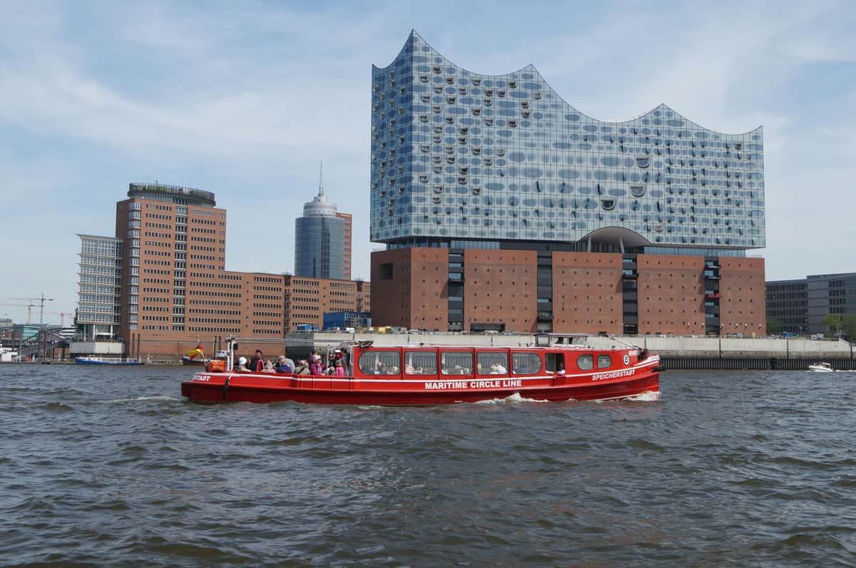 Visite du port de Hambourg sur un bateau à arrêts multiples Un bateau rouge nommé "Maritime Circle Line" avec des passagers devant des bâtiments modernes et en brique au bord de l'eau.