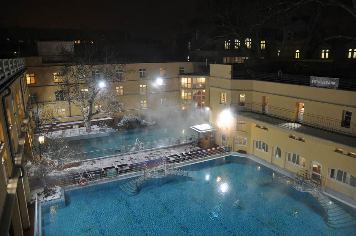 Budapest Card: 30+ Attractions with Szt. Lukacs Thermal Bath and Pool An outdoor pool area at night, with steam rising from water, surrounded by buildings with lit windows and some snow on the ground.