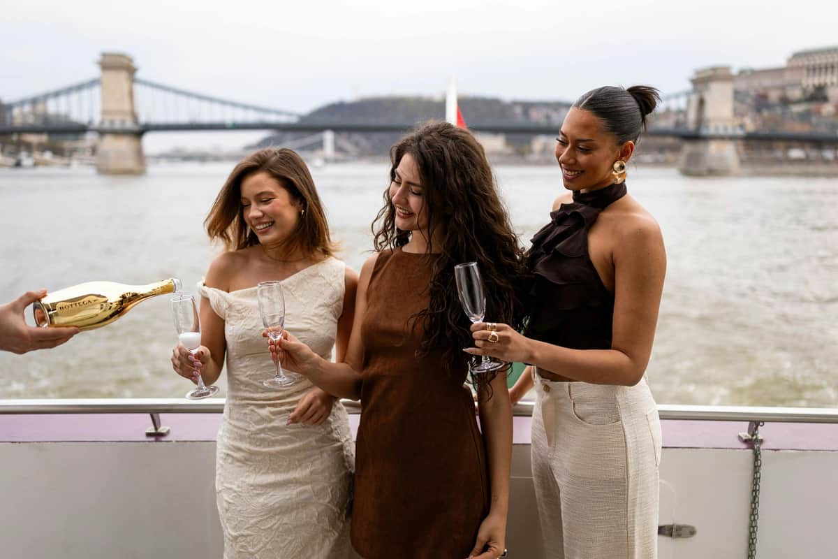 Budapest: Drinks Cruise Two women smiling, holding champagne glasses on a boat, with a bridge and river in the background.
