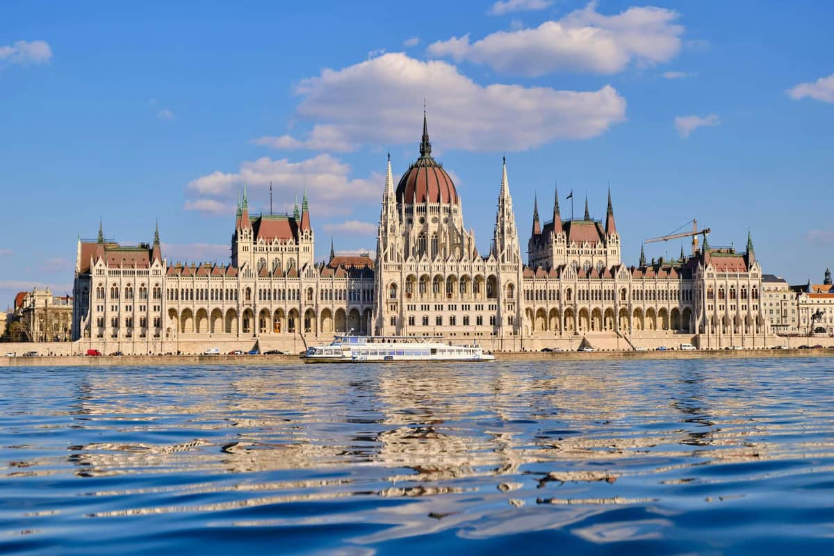 Budapest: Hop-on Hop-off Sightseeing Cruise + Audio Guide + 1 Drink A grand historical building with a dome and spires by a river under a partly cloudy sky, with a boat in the foreground.