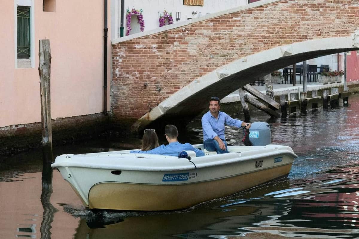 Chioggia : Visite guidée en bateau Un homme conduit un petit bateau avec deux passagers sous un pont en briques dans un canal. Des fleurs ornent le bâtiment voisin.