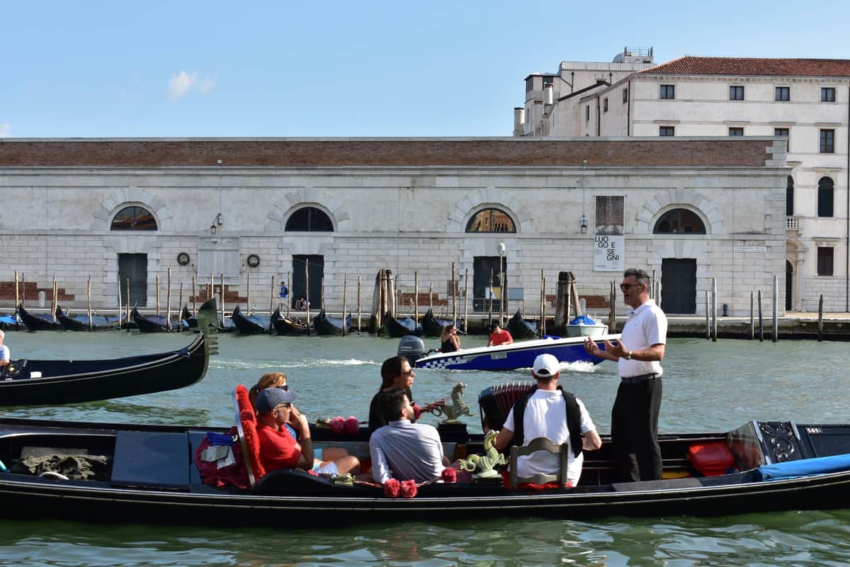 Venice: Grand Canal Evening Gondola Ride + Gallery Access + VR Experience People ride gondolas on a canal in front of a white, historic building with arched windows and doors.