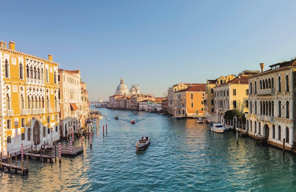 Venice Grand Canal: Guided Boat Tour for Small Group A canal in Venice with boats, lined with buildings on both sides, under a clear, blue sky. The domed structure is visible in the background.