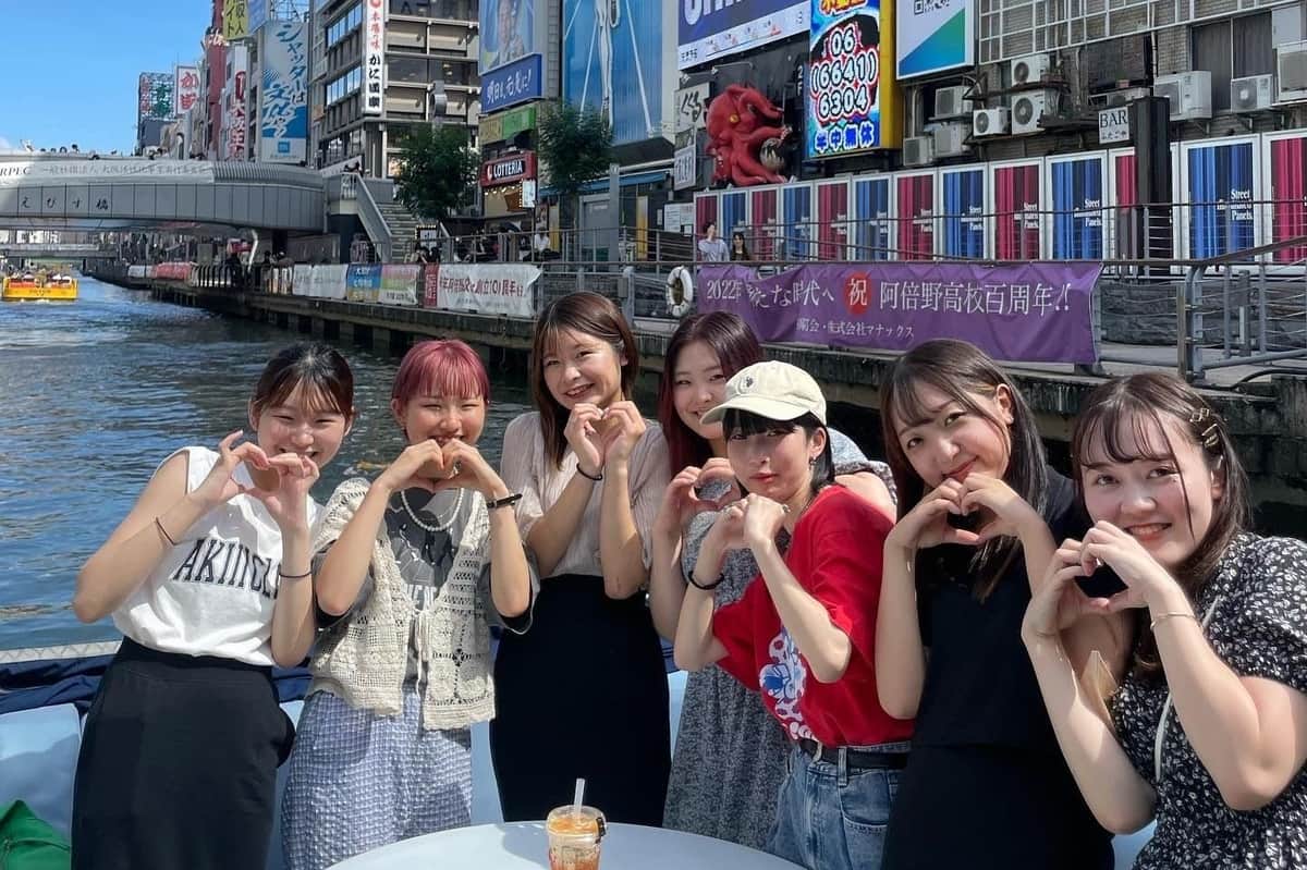 Osaka : Croisière en petit groupe sur la rivière Dotonbori Un groupe de sept femmes posant en plein air près d'un canal, faisant des gestes de la main et du cœur, avec des bâtiments urbains colorés en arrière-plan.