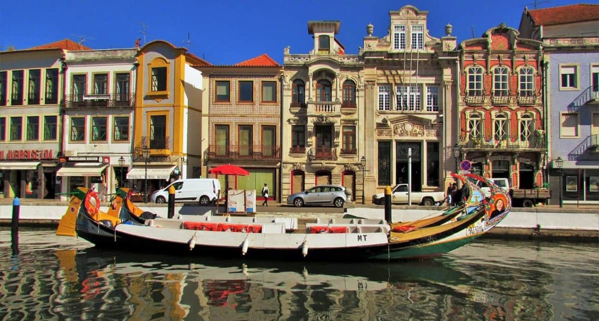 Aveiro: Half Day Tour from Porto with Boat Ride A small boat docked by a canal, in front of colorful, ornate buildings and cars under a clear blue sky.