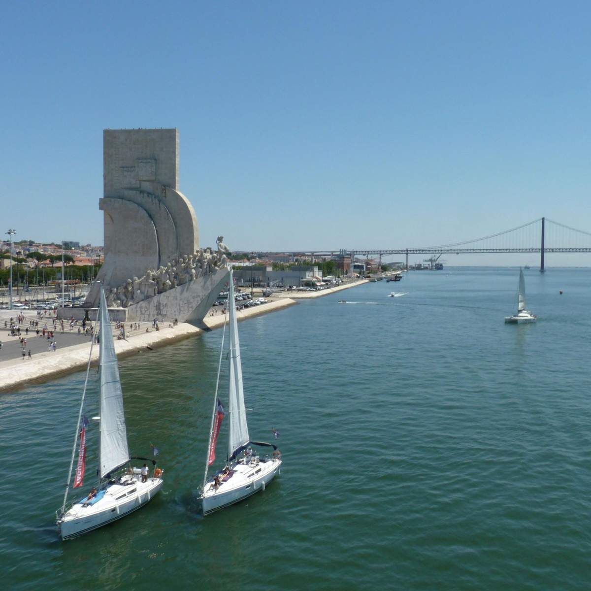 Lisbonne : 1 heure de navigation matinale à partir de Belém Deux voiliers sur une rivière près d'un grand monument en pierre et d'un long pont suspendu sous un ciel bleu clair.