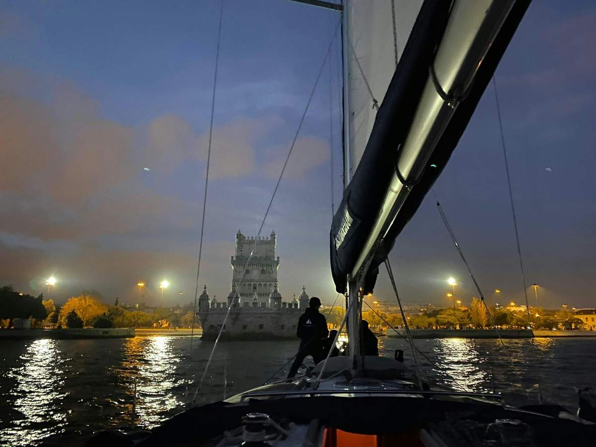 Lisbon: 2-Hour Night Sailing Tour from Belém A person sailing a boat towards a lit-up tower at dusk, with city lights in the background and calm water reflecting the lights.