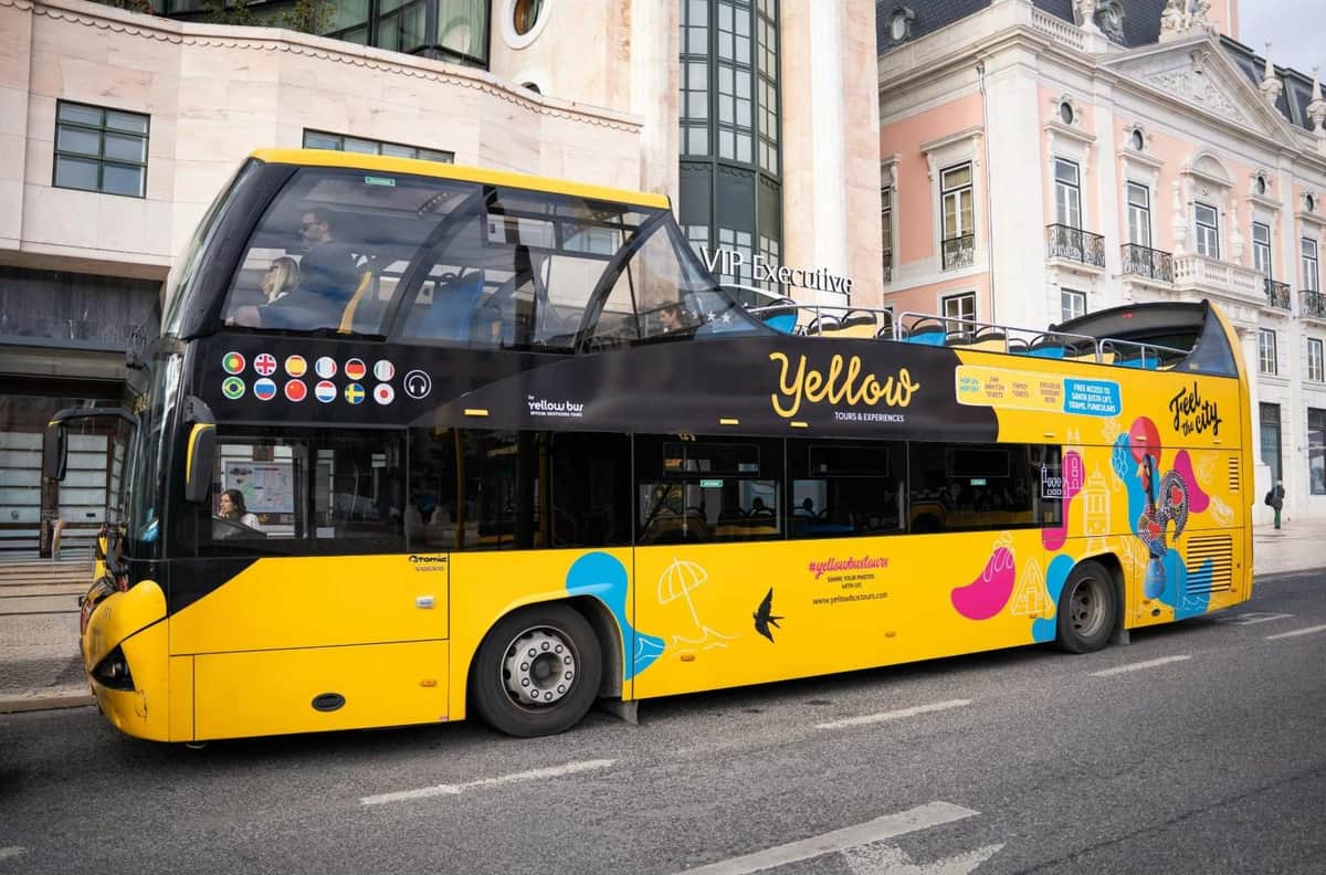Lisbonne : Bus Hop-on Hop-off valable 48 heures + croisière fluviale + visite guidée à pied de Belém Un bus touristique à impériale jaune vif et noir, orné de motifs promotionnels, stationné devant un bâtiment dans une rue de la ville.