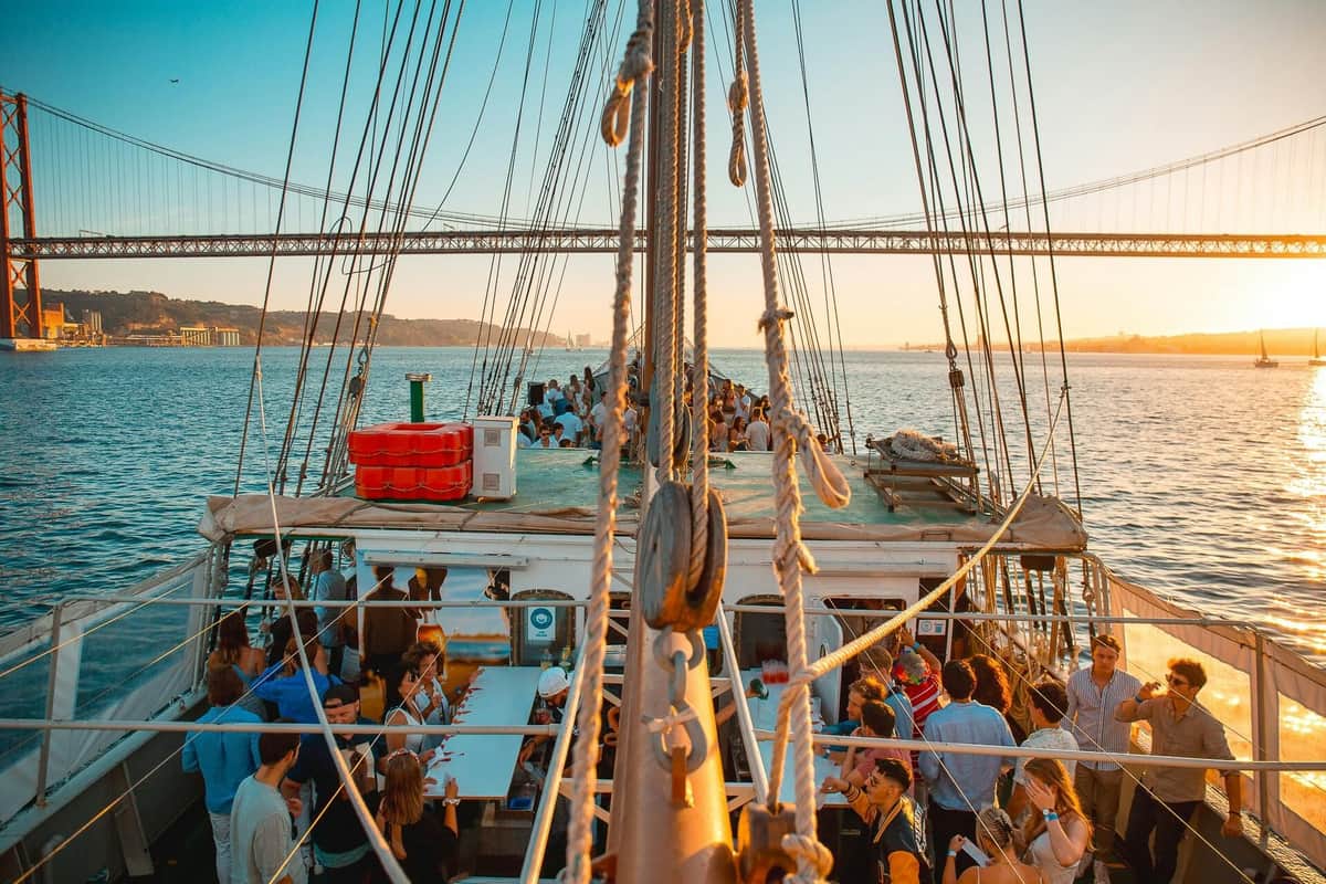 Lisbon: Sunset Boat Party + Live Music + Nightclub Entry People gather on a boat at sunset, with a suspension bridge in the background and sailboats seen on the water.