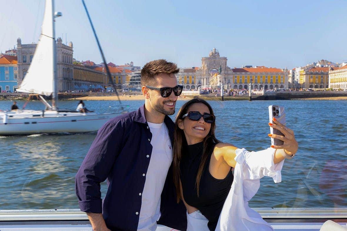 Lisbon: Tagus River Sightseeing Cruise A smiling couple takes a selfie by a waterfront with a cityscape featuring a historic building and a sailboat in the background.