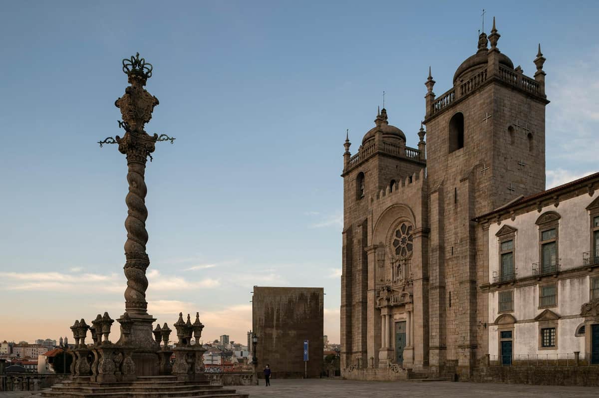 Porto Explorer Pass A historic stone cathedral with towers and intricate details stands next to a twisted column monument under a blue sky.