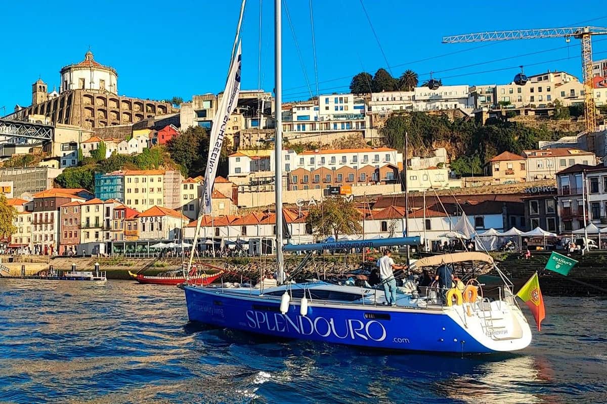 Porto: Morning or Sunset Douro River Cruise + 1 Drink A blue sailboat named Splendouro moored near a vibrant riverside town with colourful buildings and hilly terrain.