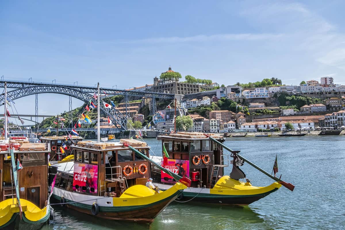 Six Bridges Cruise from Cais da Ribeira Two colorful boats on a river with a cityscape, bridge, and historic buildings in the background under a clear sky.