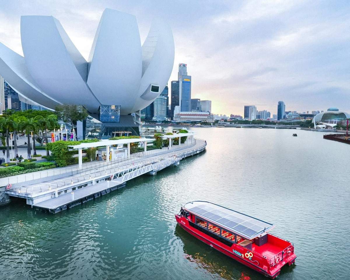 Passeio de barco da Water B: Passeio Gastronómico «Heritage» Um barco vermelho flutua numa baía, junto a um edifício branco futurista e ao horizonte da cidade, ao pôr-do-sol.